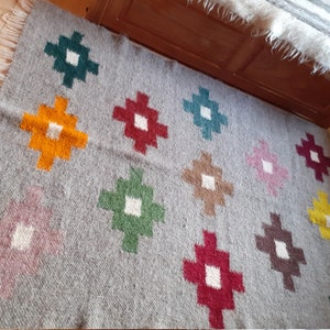 Puede incluir: Una alfombra de lana gris con un patr&oacute;n geom&eacute;trico de cuadrados coloridos. Los cuadrados est&aacute;n dispuestos en un patr&oacute;n de cuadr&iacute;cula y son en tonos de rojo, verde, amarillo, marr&oacute;n y rosa.