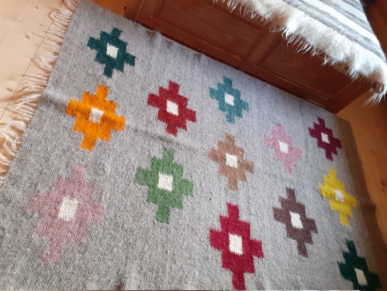 Puede incluir: Una alfombra de lana gris con un patr&oacute;n geom&eacute;trico de cuadrados coloridos. Los cuadrados est&aacute;n dispuestos en un patr&oacute;n de cuadr&iacute;cula y son en tonos de rojo, verde, amarillo, marr&oacute;n y rosa.