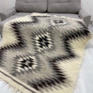 May include: A woven blanket with a geometric pattern in shades of white, gray, and black. The blanket has a fringed edge and is draped over a gray sofa. The design features a repeating diamond pattern.