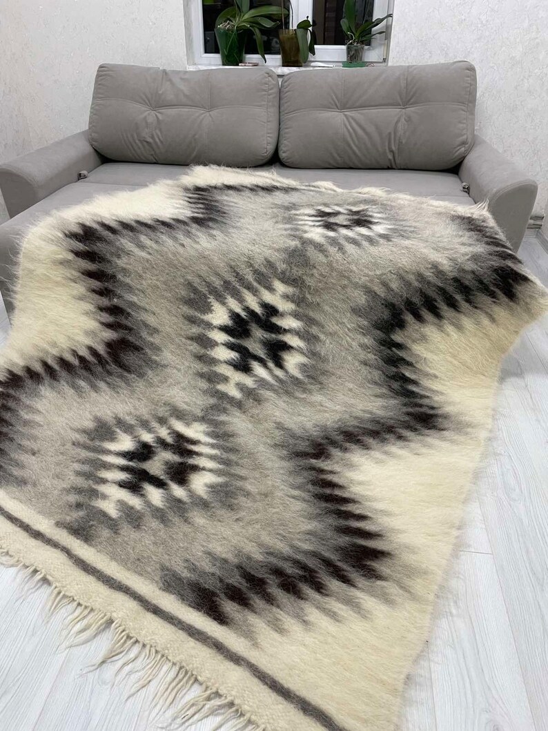 May include: A woven blanket with a geometric pattern in shades of white, gray, and black. The blanket has a fringed edge and is draped over a gray sofa. The design features a repeating diamond pattern.