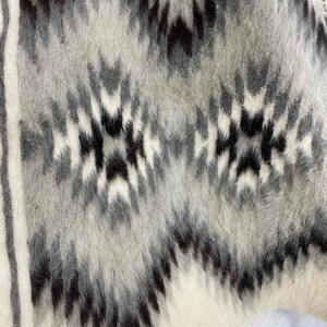 May include: Close-up of a textured, woven textile with a geometric pattern. The design features a repeating motif of black, gray, and white shapes. The fabric has a soft, fuzzy appearance, suggesting a warm, cozy material. The overall aesthetic is rustic and modern.
