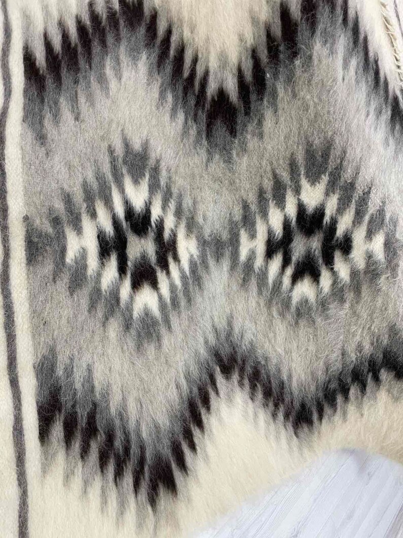 May include: Close-up of a textured, woven textile with a geometric pattern. The design features a repeating motif of black, gray, and white shapes. The fabric has a soft, fuzzy appearance, suggesting a warm, cozy material. The overall aesthetic is rustic and modern.