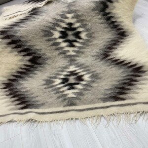 May include: A rectangular, woven rug with a geometric pattern in shades of gray, black, and cream. The rug features a central diamond design and fringed edges. The texture appears soft and plush, suitable for a living space.