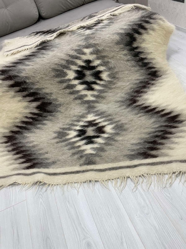 May include: A rectangular, woven rug with a geometric pattern in shades of gray, black, and cream. The rug features a central diamond design and fringed edges. The texture appears soft and plush, suitable for a living space.
