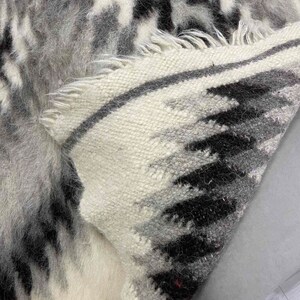 May include: A close-up of a woven rug with a pattern of black, gray, and white. The rug has a textured, shaggy appearance with fringed edges. The design features a mix of geometric and abstract shapes.