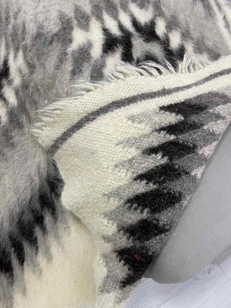 May include: A close-up of a woven rug with a pattern of black, gray, and white. The rug has a textured, shaggy appearance with fringed edges. The design features a mix of geometric and abstract shapes.