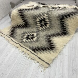 May include: A rectangular, woven blanket with a geometric pattern in shades of gray, black, and cream. The blanket has a fringed edge and is draped over a gray sofa. The design features diamond and zigzag shapes.