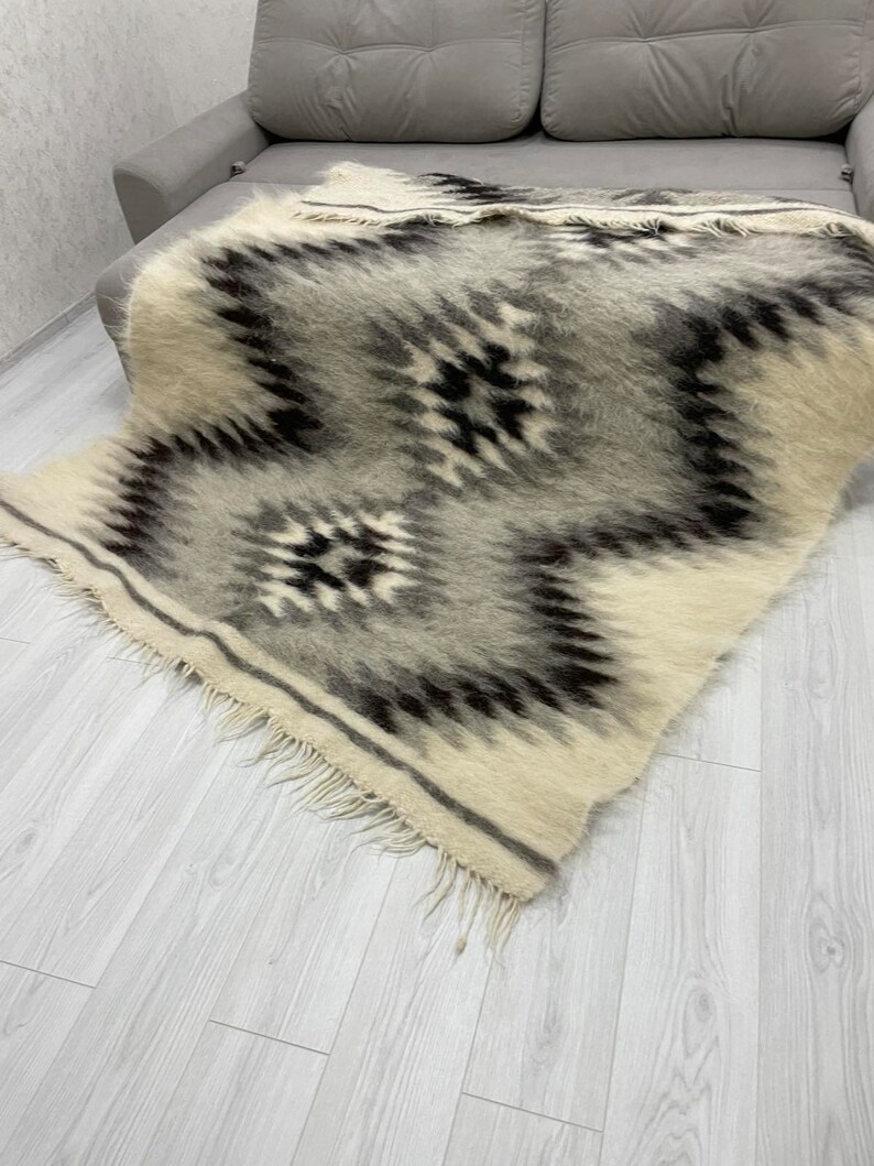 May include: A rectangular, woven blanket with a geometric pattern in shades of gray, black, and cream. The blanket has a fringed edge and is draped over a gray sofa. The design features diamond and zigzag shapes.