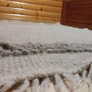 May include: A light gray, textured rug with a darker gray stripe and fringe. The rug is made of a soft, woven material and is placed on a wooden floor. The rug is near a wooden bed frame.