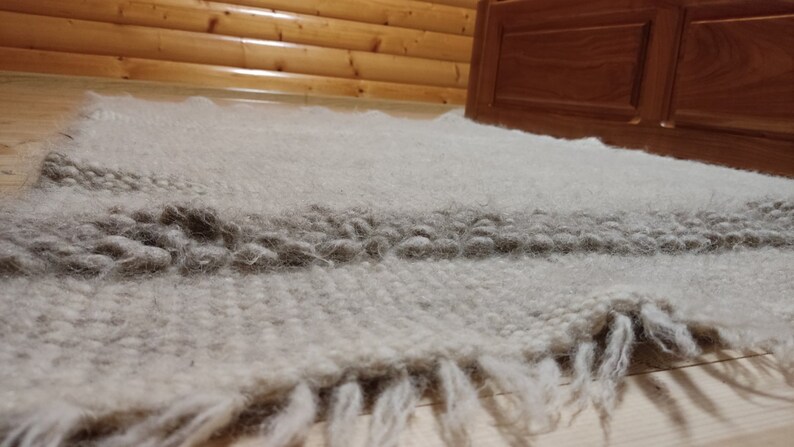 May include: A light gray, textured rug with a darker gray stripe and fringe. The rug is made of a soft, woven material and is placed on a wooden floor. The rug is near a wooden bed frame.