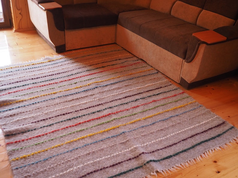 May include: A grey and white striped rug with colourful accents. The rug has a fringed border and is made of a soft, textured material.