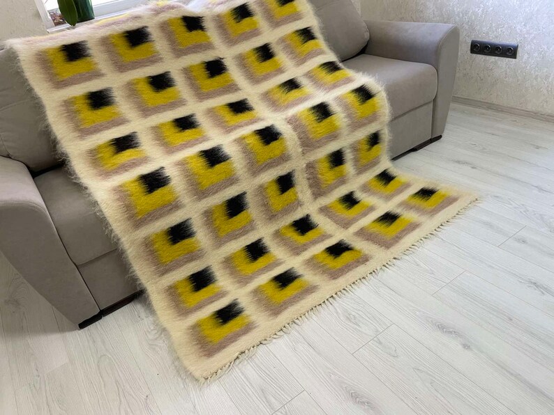 May include: A white, yellow and black patterned throw blanket with a fringe border. The blanket is draped over a grey sofa.