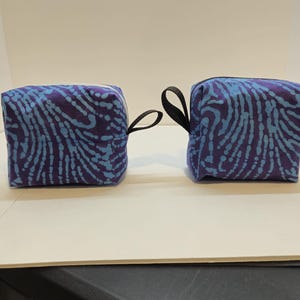 May include: Two small, square, zippered pouches with a black carrying strap. The pouches are made of a dark blue fabric with a light blue and white swirl pattern.