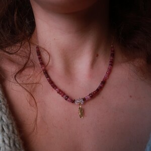 May include: A necklace featuring pink and red gemstone beads. It has a small, gold-coloured feather pendant. The necklace is worn around the neck.