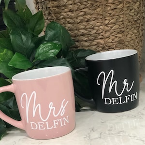 Pode incluir: Duas canecas de café de cerâmica, uma rosa e outra preta, com letras brancas. A caneca rosa diz "Sra. Delfin" e a caneca preta diz "Sr. Delfin".