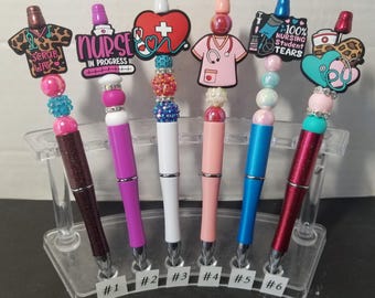 May include: Six decorative pens with nurse-themed charms. Each pen has a different color barrel and charm, including a stethoscope, scrubs, and the text "Nurse In Progress" and "100% Nursing Student Tears".