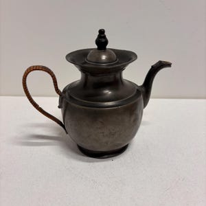 May include: Antique metal teapot with a dark finish. The teapot has a curved spout, a woven handle, and a lid with a black knob. The teapot is likely made of pewter or a similar metal. The teapot is sitting on a white surface.