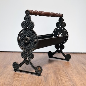 May include: An antique black metal magazine rack with ornate, circular side designs and a dark wooden handle. The rack has a rectangular basket for holding magazines and decorative scrollwork on the legs.