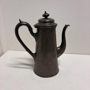 Op de afbeelding: Een donkergrijze metalen koffiepot met een gebogen handvat en een tuit. De pot heeft een deksel met een kleine knop bovenop. Het handvat is aan de zijkant bevestigd en buigt omhoog. De tuit is gebogen.