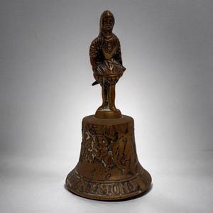 May include: Bronze bell with a figure of a person in armor holding a drum. The bell has an embossed design of figures and the word "ZHEMONY" around the base. The bell is a vintage item.