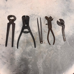 Op de afbeelding: Een assortiment vintage metalen gereedschappen, waaronder tangen, kniptangen en een sleutel, gerangschikt op een getextureerd oppervlak. De gereedschappen zijn in verschillende tinten donkergrijs en bruin, met tekenen van ouderdom en gebruik. De gereedschappen worden waarschijnlijk gebruikt voor constructie of reparatie.