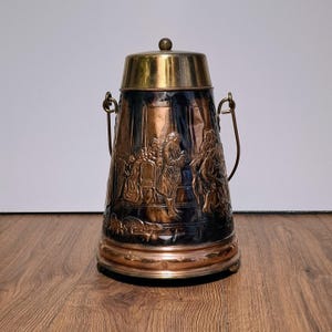 May include: A copper and brass ice bucket with a lid and handle. The bucket features a detailed relief scene of people in period clothing. The top and lid are brass, while the body is copper with a dark patina.