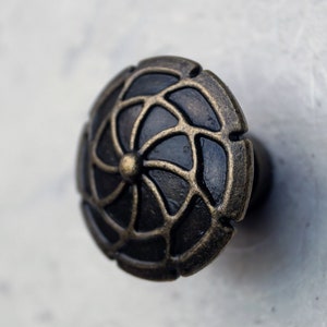 Puede incluir: Un pomo de mueble redondo con un centro negro mate oscuro y una superposición de metal en tono latón antiguo en relieve. La superposición muestra un patrón simétrico de pétalos giratorios que irradian desde un punto central.