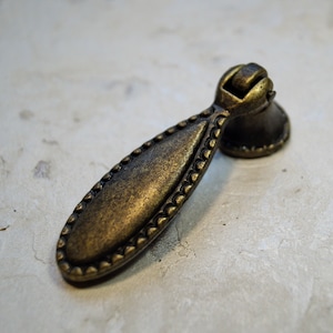 May include: A gold-toned metal cabinet pull with a decorative beaded edge and a pointed oval shape.