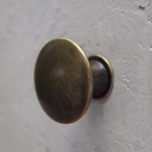 May include: Close-up of an antique brass cabinet knob. The knob has an oval shape with a slightly textured surface and a dark, aged finish. The knob is mounted on a light grey surface.