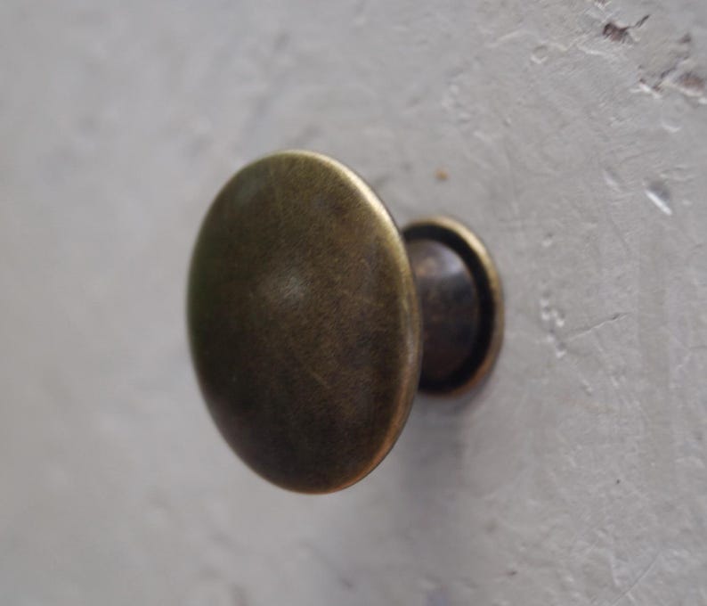May include: Close-up of an antique brass cabinet knob. The knob has an oval shape with a slightly textured surface and a dark, aged finish. The knob is mounted on a light grey surface.