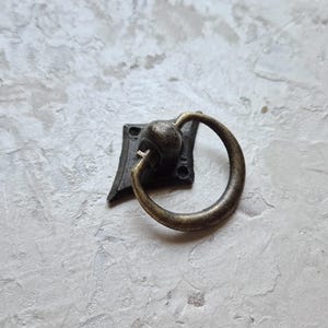 May include: Antique bronze-finished drawer pull with a ring handle. The pull is mounted on a square backplate with decorative edges. The ring handle is attached to a round base.