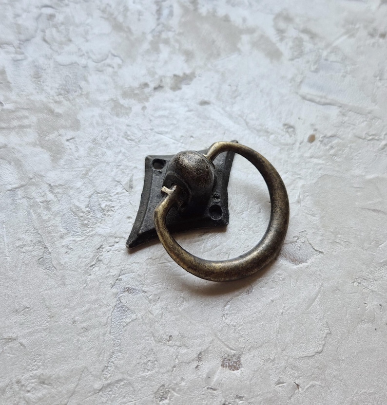 May include: Antique bronze-finished drawer pull with a ring handle. The pull is mounted on a square backplate with decorative edges. The ring handle is attached to a round base.