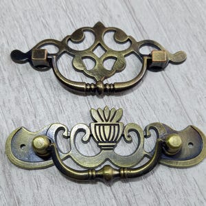 Pode incluir: Duas puxadores de gaveta de metal adornadas com um design floral decorativo. A puxador superior é um simples laço com uma maçaneta quadrada em cada extremidade. A puxador inferior é um design mais elaborado com um motivo floral central e uma maçaneta redonda em cada extremidade.