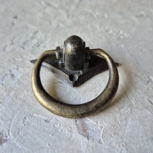 May include: Antique brass drawer pull with a ring handle. The pull has a decorative backplate with a central mounting point. The ring handle is circular and attached to the backplate. The finish has a distressed appearance.