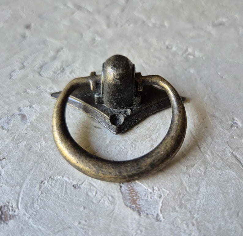May include: Antique brass drawer pull with a ring handle. The pull has a decorative backplate with a central mounting point. The ring handle is circular and attached to the backplate. The finish has a distressed appearance.