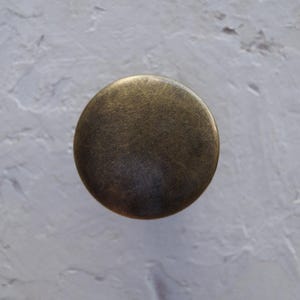 May include: A circular cabinet knob in an antique brass finish. The knob has a slightly curved profile and a distressed appearance, suggesting a vintage aesthetic. Suitable for drawers, cabinets, or other furniture, it measures approximately 3 cm in diameter.