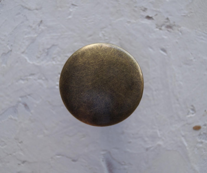 May include: A circular cabinet knob in an antique brass finish. The knob has a slightly curved profile and a distressed appearance, suggesting a vintage aesthetic. Suitable for drawers, cabinets, or other furniture, it measures approximately 3 cm in diameter.