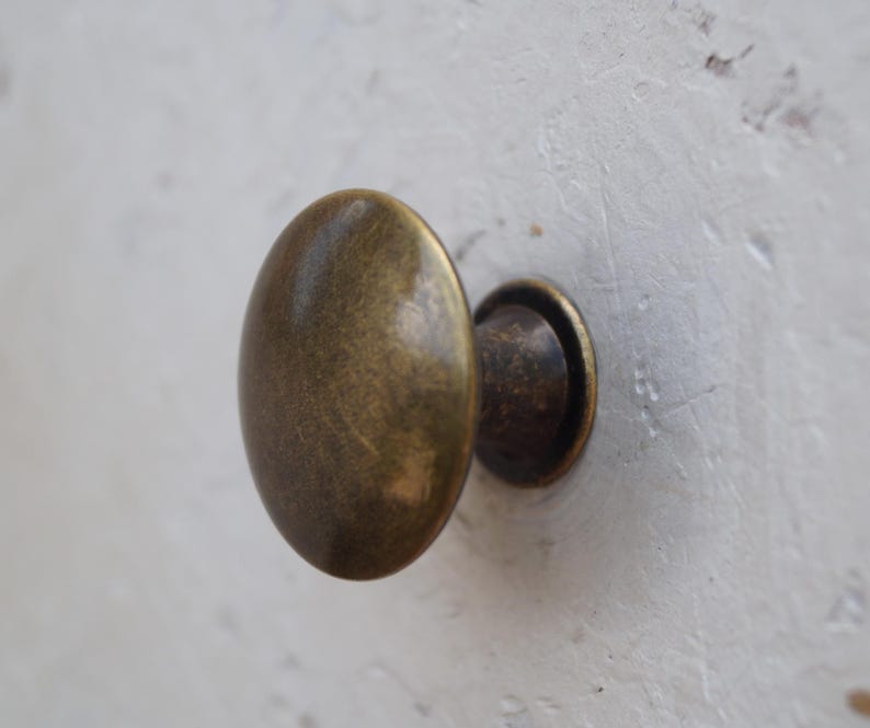 May include: An oval-shaped, antique brass cabinet knob. The knob has a rounded, smooth surface and a small, circular base. The hardware is mounted on a white, textured surface. The knob is a warm, metallic colour.