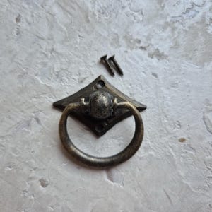 May include: Antique bronze drawer pull with a diamond-shaped backplate and a rounded ring handle. Two small screws are included. The hardware has a distressed finish, suitable for cabinets, drawers, or furniture restoration projects.