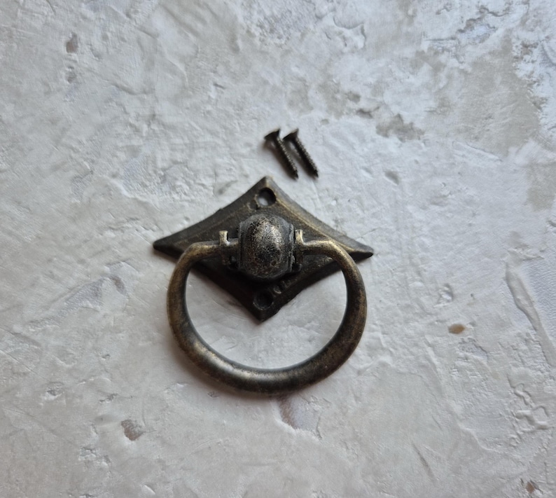 May include: Antique bronze drawer pull with a diamond-shaped backplate and a rounded ring handle. Two small screws are included. The hardware has a distressed finish, suitable for cabinets, drawers, or furniture restoration projects.