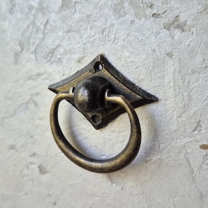 May include: Antique brass drawer pull with a diamond-shaped backplate and a circular handle. The hardware has a vintage aesthetic, perfect for adding a touch of classic style to furniture or cabinetry. The pull is mounted on a textured, light-colored surface.