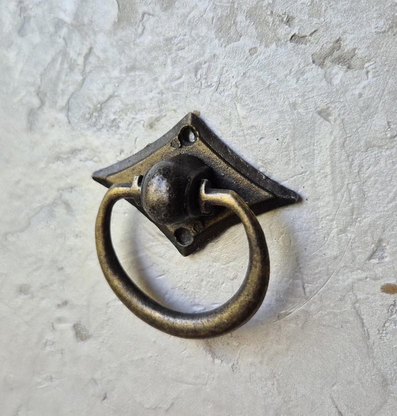 May include: Antique brass drawer pull with a diamond-shaped backplate and a circular handle. The hardware has a vintage aesthetic, perfect for adding a touch of classic style to furniture or cabinetry. The pull is mounted on a textured, light-colored surface.