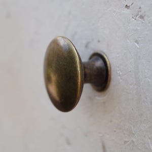 May include: Close-up of a bronze cabinet knob with an oval head. The knob is fixed to a textured, off-white surface, possibly a door or drawer. The metal has a slightly aged appearance, suggesting a vintage style.