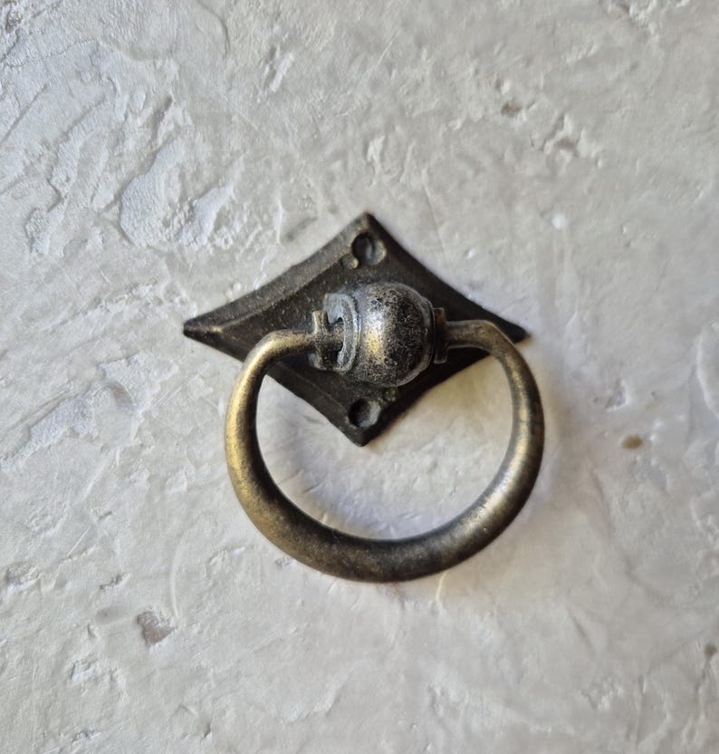 May include: Antique bronze-finished drawer pull with a diamond-shaped backplate and a rounded ring handle. The hardware has a distressed finish, adding a vintage aesthetic. Ideal for cabinets, drawers, or furniture restoration projects.