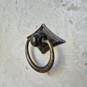 May include: Antique bronze-finished drawer pull with a diamond-shaped backplate and a circular ring handle. The hardware is mounted on a textured, light-colored surface. The pull is designed for cabinets, drawers, or furniture.
