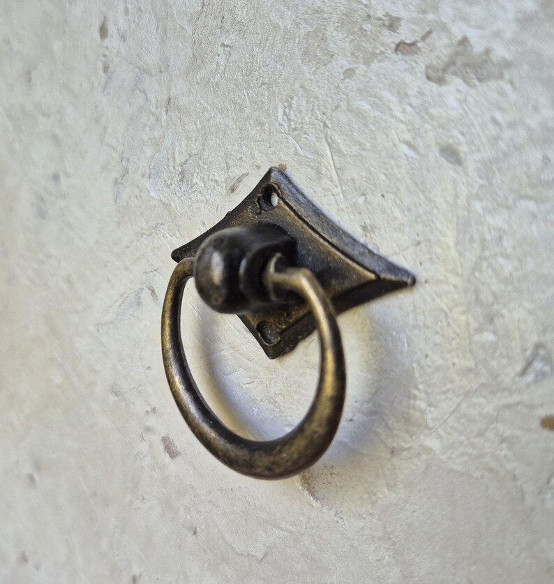 May include: Antique bronze-finished drawer pull with a diamond-shaped backplate and a circular ring handle. The hardware is mounted on a textured, light-colored surface. The pull is designed for cabinets, drawers, or furniture.