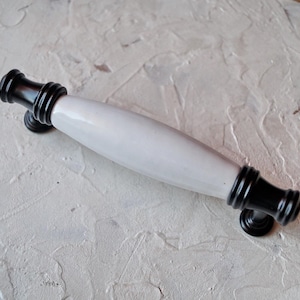 May include: A cabinet pull with a white ceramic center and black metal ends. The handle has a cylindrical shape with decorative ridges near the ends. The hardware is designed for easy installation and adds a touch of elegance.