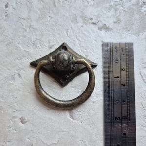 May include: Antique bronze drawer pull with a diamond-shaped backplate and a circular handle. The pull is approximately 1 inch wide. The ruler shows measurements in inches and centimeters.