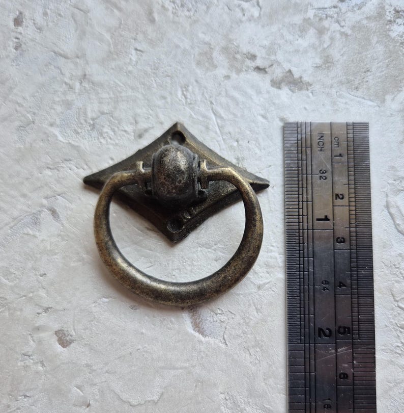 May include: Antique bronze drawer pull with a diamond-shaped backplate and a circular handle. The pull is approximately 1 inch wide. The ruler shows measurements in inches and centimeters.