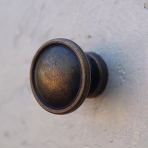 Puede incluir: Un pomo de armario redondo de estilo antiguo con un acabado en bronce oscuro. El pomo tiene un diseño circular elevado y una superficie ligeramente texturizada.